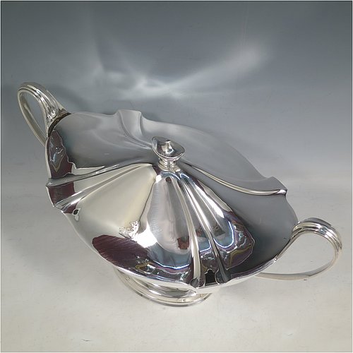 An Antique Edwardian Sterling silver Neoclassical style soup tureen, having a pull-off lid with an urn-shaped finial and cut-out slot for a ladle, with hand-chased shoulders, applied reeded borders, two reeded scroll side-handles, and all sitting on a pedestal foot. Made by Goldsmiths & Silversmiths of London in 1902. The dimensions of this fine hand-made antique silver soup tureen and cover are height 23 cms (9 inches), length including handles 38 cms (15 inches), width 20 cms (8 inches), and it weighs approx. 1,615 (52 troy ounces). Please note that this item is crested on lid and body.