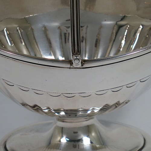 An Antique Georgian Sterling Silver sugar basket, having an oval bellied body with hand-chased half-fluting, with applied reeded borders, a hinged and reeded swing-handle, and sitting on a pedestal foot. Made by Peter & Anne Bateman of London in 1791. The dimensions of this fine hand-made antique silver sugar basket are height (inc. handle) 15 cms (6 inches), length 14 cms (5.5 inches), and it weighs approx. 198g (6.4 troy ounces).
