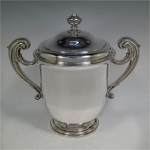 A very handsome and heavy Antique Victorian Sterling Silver trophy cup and cover, in a George II style, having a plain round body, two flying scroll side-handles with anthemion leaf thumb-pieces, a pull-off lid with cast finial, and all sitting on a stepped pedestal foot. Made by William Barnard of London in 1895. The dimensions of this fine hand-made antique silver trophy cup and cover are height 27 cms (10.5 inches), spread across arms 30.5 cms (12 inches), and it weighs approx. 1,787g (57.6 troy ounces).   