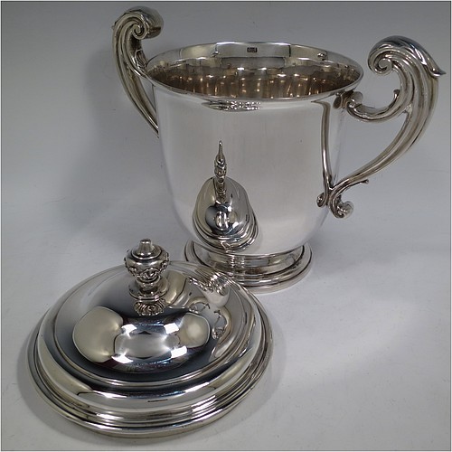 A very handsome and heavy Antique Victorian Sterling Silver trophy cup and cover, in a George II style, having a plain round body, two flying scroll side-handles with anthemion leaf thumb-pieces, a pull-off lid with cast finial, and all sitting on a stepped pedestal foot. Made by William Barnard of London in 1895. The dimensions of this fine hand-made antique silver trophy cup and cover are height 27 cms (10.5 inches), spread across arms 30.5 cms (12 inches), and it weighs approx. 1,787g (57.6 troy ounces).