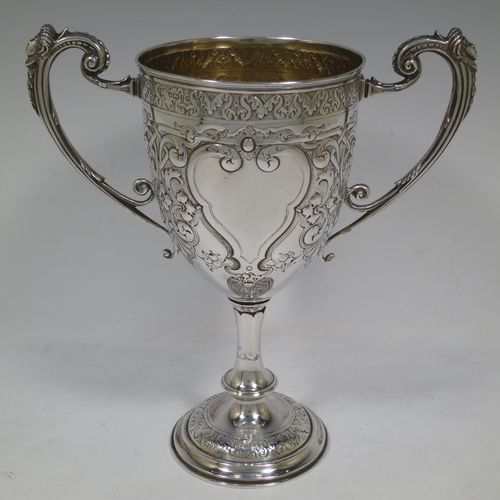 A very beautiful Antique Victorian Sterling Silver trophy cup, having a round body with tapering sides all hand-chased with floral and scroll decoration surrounding vacant cartouches either side, two cast scroll handles with figural thumb-pieces, a gold-gilt interior, and all sitting on a pedestal foot. Made by John Edward Bingham and Charles Henry Bingham of Sheffield in 1876. The dimensions of this fine hand-made antique silver trophy cup are height 24 cms (9.5 inches), spread across arms 22 cms (8.75 inches), and it weighs approx. 620g (20 troy ounces).   