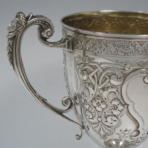 A very beautiful Antique Victorian Sterling Silver trophy cup, having a round body with tapering sides all hand-chased with floral and scroll decoration surrounding vacant cartouches either side, two cast scroll handles with figural thumb-pieces, a gold-gilt interior, and all sitting on a pedestal foot. Made by John Edward Bingham and Charles Henry Bingham of Sheffield in 1876. The dimensions of this fine hand-made antique silver trophy cup are height 24 cms (9.5 inches), spread across arms 22 cms (8.75 inches), and it weighs approx. 620g (20 troy ounces).