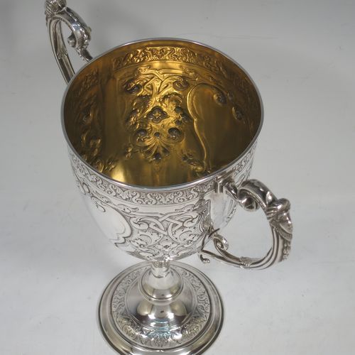 A very beautiful Antique Victorian Sterling Silver trophy cup, having a round body with tapering sides all hand-chased with floral and scroll decoration surrounding vacant cartouches either side, two cast scroll handles with figural thumb-pieces, a gold-gilt interior, and all sitting on a pedestal foot. Made by John Edward Bingham and Charles Henry Bingham of Sheffield in 1876. The dimensions of this fine hand-made antique silver trophy cup are height 24 cms (9.5 inches), spread across arms 22 cms (8.75 inches), and it weighs approx. 620g (20 troy ounces).