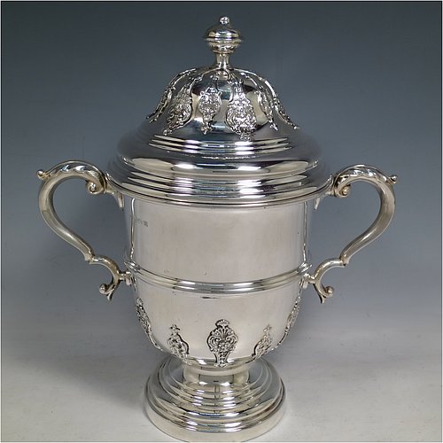 A Sterling Silver trophy cup and cover, in a George II style, having a round body with applied floral strap-work decoration, two scroll side-handles with anthemion leaf thumb-pieces, a central applied girdle band, a pull-off lid with matching applied strap-work and a cast round finial, and all sitting on a stepped pedestal foot. Made by George Bryan & Co., of Birmingham in 1936. The dimensions of this fine hand-made silver trophy cup and cover are height 30 cms (11.75 inches), spread across arms 26.5 cms (10.5 inches), and it weighs approx. 1,132g (36.5 troy ounces).    