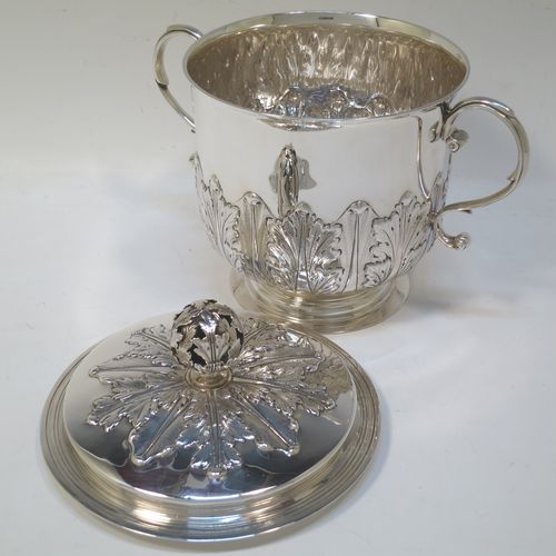 A very handsome Antique Sterling Silver trophy cup and cover, in the shape of a lidded Porringer, having a round body with hand-chased Anthemion leaf decoration, with two scroll side-handles, a lift-off cover with matching decoration and a pierced floral finial, and all sitting on a stepped pedestal foot. This elegant antique silver trophy cup and cover was made by Charles Stuart Harris of London in 1925. The dimensions of this fine hand-made antique silver trophy cup and cover are height 18 cms (7 inches), spread across handles 21 cms (8.25 inches), and it weighs approx. 748g (24 troy ounces).
