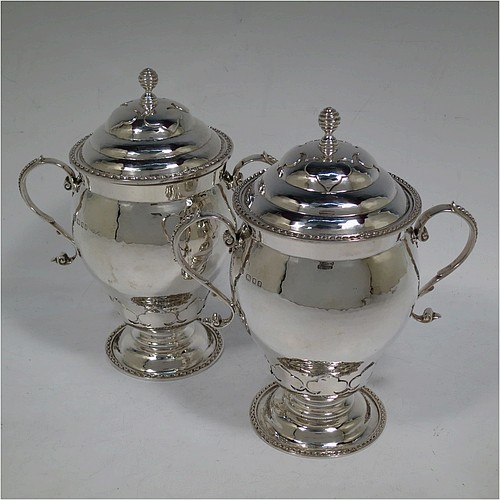 A very unusual pair of Sterling Silver trophy cups and covers, in an Arts and Crafts style, having round bellied bodies with hand-hammered decoration, applied zigzag borders and cut-card work, two side scroll handles with beaded thumb-pieces, removable covers with spiral finials, and all sitting on stepped pedestal feet. Made by D. and J. Wellby of London in 1931. The dimensions of these fine hand-made silver trophy cups and covers are height 17.5 cms (7 inches), spread across arms 15 cms (6 inches), and they weigh a total of  approx. 825g (26.6 troy ounces).   