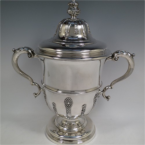 A very handsome Antique Edwardian Sterling Silver trophy cup and cover, in a George II style, having a round body with applied floral strap-work decoration, two scroll side-handles with anthemion leaf thumb-pieces, a central applied girdle band, a pull-off lid with matching applied strap-work and a cast round floral finial, and all sitting on a stepped pedestal foot. Made by Walker and Hall of Sheffield in 1909. The dimensions of this fine hand-made antique silver trophy cup and cover are height 37 cms (14.5 inches), spread across arms 33 cms (13 inches), and it weighs approx. 1,570g (51 troy ounces).    