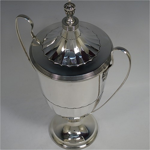 A very handsome Sterling Silver trophy cup and cover in a George III neoclassical style, having a round body with tapering sides and half-fluted decoration, with hand-chased reeded borders, two looped side-handles, an original cover with matching decoration and cast urn finial, and all sitting on a pedestal foot. Made by Robert Pringle of London in 1923. The dimensions of this fine hand-made silver trophy cup and cover are height 33 cms (13 inches), spread across arms 21 cms (8.25 inches), and it weighs approx. 879g (28 troy ounces).