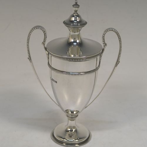 A small but very elegant Antique Sterling Silver trophy cup and cover in a George III neoclassical style, having a round plain body with tapering sides, with applied bead-edged borders, two looped side-handles, an original cover with matching decoration and cast urn finial, and all sitting on a pedestal foot. This handsome silver trophy cup was made by William Hutton of Sheffield in 1912. The dimensions of this fine hand-made silver trophy cup and cover are height 20 cms (8 inches), spread across arms 12.5 cms (5 inches), and it weighs approx. 280g (9 troy ounces).   