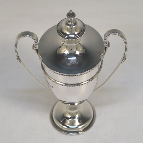 A small but very elegant Antique Sterling Silver trophy cup and cover in a George III neoclassical style, having a round plain body with tapering sides, with applied bead-edged borders, two looped side-handles, an original cover with matching decoration and cast urn finial, and all sitting on a pedestal foot. This handsome silver trophy cup was made by William Hutton of Sheffield in 1912. The dimensions of this fine hand-made silver trophy cup and cover are height 20 cms (8 inches), spread across arms 12.5 cms (5 inches), and it weighs approx. 280g (9 troy ounces).   