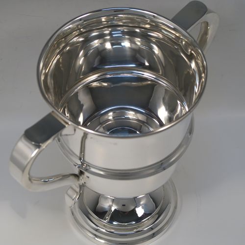 A very handsome Sterling Silver trophy cup in a George III neoclassical style, having a round plain body with tapering sides, an applied central reeded band, two plain scroll side-handles, and all sitting on a stepped pedestal foot. This elegant silver trophy cup was made by Carrington and Co., of Birmingham in 1931. The dimensions of this fine hand-made silver trophy cup are height 21 cms (8.25 inches), spread across arms 23.5 cms (9.25 inches), and it weighs approx. 715g (23 troy ounces).  