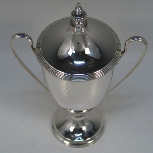 A very handsome Antique Edwardian Sterling Silver trophy cup and cover in a George III neoclassical style, having a round plain body with tapering sides, with hand-chased reeded borders, two looped side-handles, an original cover with matching decoration and cast knop finial, and all sitting on a pedestal foot. This beautiful silver trophy cup was made by Elkington and Co., of Birmingham in 1908. The dimensions of this fine hand-made silver trophy cup and cover are height 24 cms (9.5 inches), spread across arms 16.5 cms (6.5 inches), and it weighs approx. 387g (12.5 troy ounces).