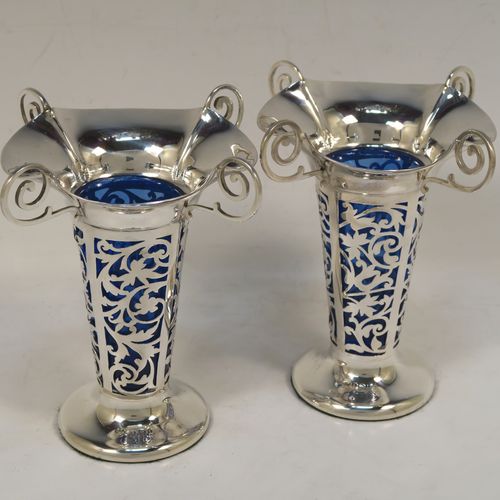A vert pretty pair of Antique Edwardian Sterling Silver vases, having hand-pierced bodies with floral decoration on round tapering sides, with removable blue-glass liners, with wavy-edge rims and four scroll handles, and all sitting on plain round pedestal feet. These beautiful antique silver vases were made by Harrison and Hipwood of Birmingham in 1910. The dimensions of this fine hand-made pair of antique silver vases are height 13 cms (5.25 inches), and diameter at top 10 cms (4 inches).   