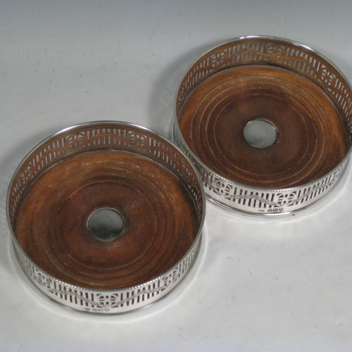 Sterling silver pair of wine bottle table coasters, having round bodies with hand-pierced decoration and engraved swags, with wood-turned bases and silver buttons, and applied beaded borders. Made by Henry Freeman Ltd., of London in 1913. The dimensions of these wine coasters are diameter 13 cms (5 inches), height 4 cms (1.5 inches).