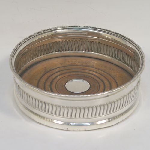 A very handsome Antique Georgian Sterling Silver wine bottle table coaster, in a Regency style, having a round bellied body with a hand-chased band of vertical fluting, an applied rope-twist border, and an oak wood-turned base with central silver button. This elegant antique silver wine bottle coaster was made in Sheffield in 1805. The dimensions of this fine hand-made silver wine bottle coaster are diameter 13 cms (5.25 inches), and height 4.5 cms (1.75 inches).   