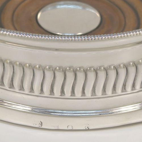 A very handsome Antique Georgian Sterling Silver wine bottle table coaster, in a Regency style, having a round bellied body with a hand-chased band of vertical fluting, an applied rope-twist border, and an oak wood-turned base with central silver button. This elegant antique silver wine bottle coaster was made in Sheffield in 1805. The dimensions of this fine hand-made silver wine bottle coaster are diameter 13 cms (5.25 inches), and height 4.5 cms (1.75 inches).   