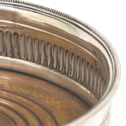 A very handsome Antique Georgian Sterling Silver wine bottle table coaster, in a Regency style, having a round bellied body with a hand-chased band of vertical fluting, an applied rope-twist border, and an oak wood-turned base with central silver button. This elegant antique silver wine bottle coaster was made in Sheffield in 1805. The dimensions of this fine hand-made silver wine bottle coaster are diameter 13 cms (5.25 inches), and height 4.5 cms (1.75 inches).   