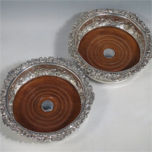 An Antique Georgian Sterling Silver pair of table wine bottle coasters, having round bellied bodies with hand-chased grape and vine decoration, and applied shell and scroll borders, and wood turned bases with silver buttons. Made S. C. Younge & Co of Sheffield in 1824. The dimensions of this fine pair of hand-made antique silver wine bottle coasters are diameter 16.5 cms (6.5 inches), and height 5 cms (2 inches).    