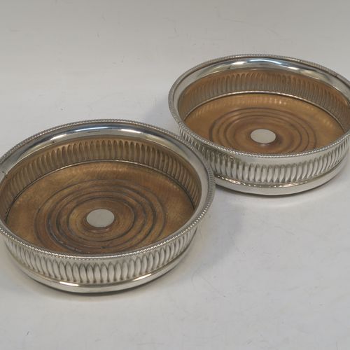 A very handsome Antique Georgian Old Sheffield plated pair of table wine coasters, in a Regency style, having round bellied bodies, with applied gadroon borders, and hand-chased fluted decoration, and with wood-turned bases and silver buttons. Made in ca. 1810. The dimensions of this fine pair of antique hand-made Old Sheffield plated wine bottle coasters are diameter 14 cms (5.5 inches), and height 4 cms (1.5 inches).