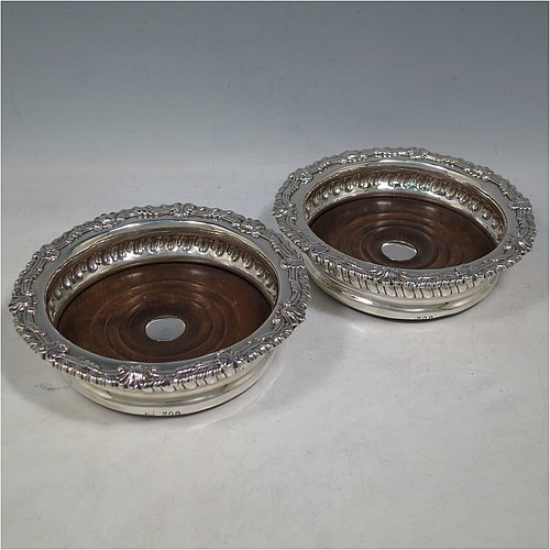 An Antique Georgian Sterling Silver pair of table wine bottle coasters, in a Regency style having round bodies with a band of hand-chased fluted decoration, applied gadroon, scroll, & shell borders, and wood turned bases with silver buttons. Made S. C. Younge & Co of Sheffield in 1815. The dimensions of this fine pair of hand-made antique silver wine bottle coasters are diameter 16 cms (6.3 inches), and height 4 cms (1.5 inches).    