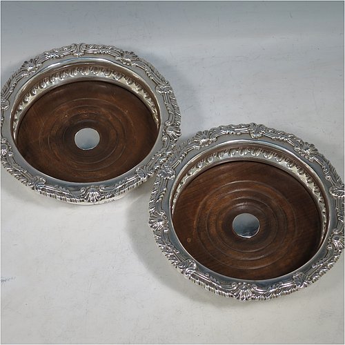An Antique Georgian Sterling Silver pair of table wine bottle coasters, in a Regency style having round bodies with a band of hand-chased fluted decoration, applied gadroon, scroll, & shell borders, and wood turned bases with silver buttons. Made S. C. Younge & Co of Sheffield in 1815. The dimensions of this fine pair of hand-made antique silver wine bottle coasters are diameter 16 cms (6.3 inches), and height 4 cms (1.5 inches).