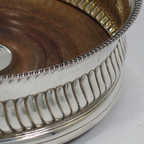 A very handsome Antique Georgian Sterling Silver wine bottle table coaster, in a Regency style, having a round bellied body with a hand-chased band of vertical fluting, an applied gadroon border, and an oak wood-turned base with central silver button. Made in London in 1808. The dimensions of this fine hand-made silver wine bottle coaster are diameter 14 cms (5.5 inches), and height 5 cms (2 inches).