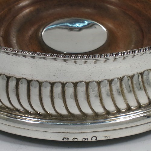 A very handsome Antique Georgian Sterling Silver wine bottle table coaster, in a Regency style, having a round bellied body with a hand-chased band of vertical fluting, an applied gadroon border, and an oak wood-turned base with central silver button. Made in London in 1808. The dimensions of this fine hand-made silver wine bottle coaster are diameter 14 cms (5.5 inches), and height 5 cms (2 inches).