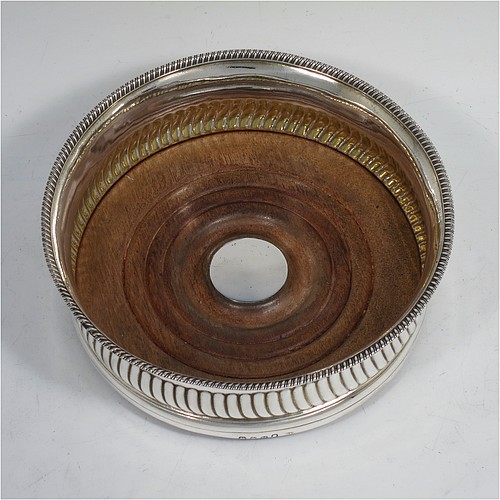 A very handsome Antique Georgian Sterling Silver wine bottle table coaster, in a Regency style, having a round bellied body with a hand-chased band of vertical fluting, an applied gadroon border, and an oak wood-turned base with central silver button. Made in London in 1808. The dimensions of this fine hand-made silver wine bottle coaster are diameter 14 cms (5.5 inches), and height 5 cms (2 inches).