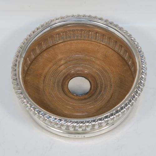 A very handsome Antique Georgian Sterling Silver wine bottle table coaster, in a Regency style, having a round bellied body with a hand-chased melon vertical fluting, an applied gadroon border, and an oak wood-turned base with central silver button. This elegant silver bottle coaster was made by Thomas and John Settle of London in 1819. The dimensions of this fine hand-made antique silver wine bottle coaster are diameter 15 cms (6 inches), and height 5 cms (2 inches).