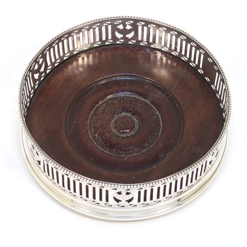 A very pretty Antique Georgian Neoclassical style Sterling Silver wine bottle table coaster, having a round hand-pierced body with geometrical and tureen style decoration, together with an applied bead-edged top border, and an wood-turned base. This beautiful antique silver wine bottle coaster was made in London in 1786. The dimensions of this fine hand-made antique silver wine bottle coaster are diameter 12 cms (4.75 inches), and height 3.5 cms (1.3 inches).
