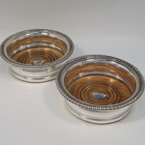 A very handsome pair of Antique Georgian Sterling Silver wine bottle table coasters, in a Georgian Regency style, having round bellied bodies with applied gadroon borders, and oak wood-turned bases with silver buttons. These elegant antique silver wine coasters were made by John and Thomas Settle of Sheffield in 1821. The dimensions of these fine hand-made antique silver wine bottle coasters are diameter 16 cms (6.3 inches), and height 5 cms (2 inches). Please note that the silver buttons are part-crested.