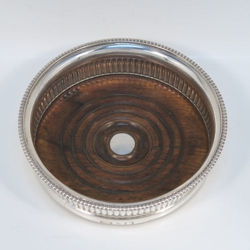 A very handsome Antique Georgian Sterling Silver wine bottle table coaster, in a Regency style, having a round bellied body with a hand-chased melon vertical fluting, an applied gadroon border, and an oak wood-turned base with central silver button. This elegant silver bottle coaster was made by John Roberts of Sheffield in 1806. The dimensions of this fine hand-made antique silver wine bottle coaster are diameter 14.5 cms (5.75 inches), and height 4 cms (1.5 inches).